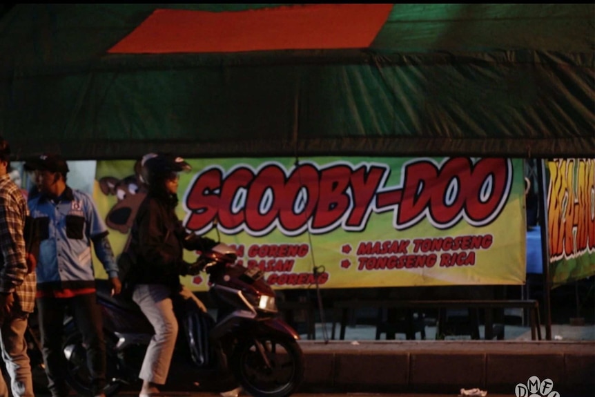 A person on a motorbike rides past a large sign saying "Scooby-doo".