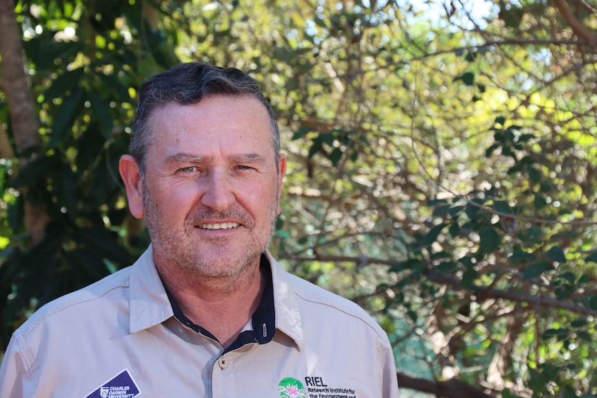 A man wearing a collared khaki shirt smiling at the camera, standing in front of trees.