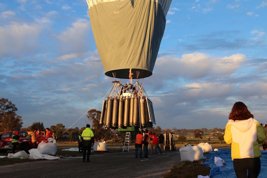 A hot-air balloon inches away from the ground with oxygen bottles on the outside of the gondola as people watch on.