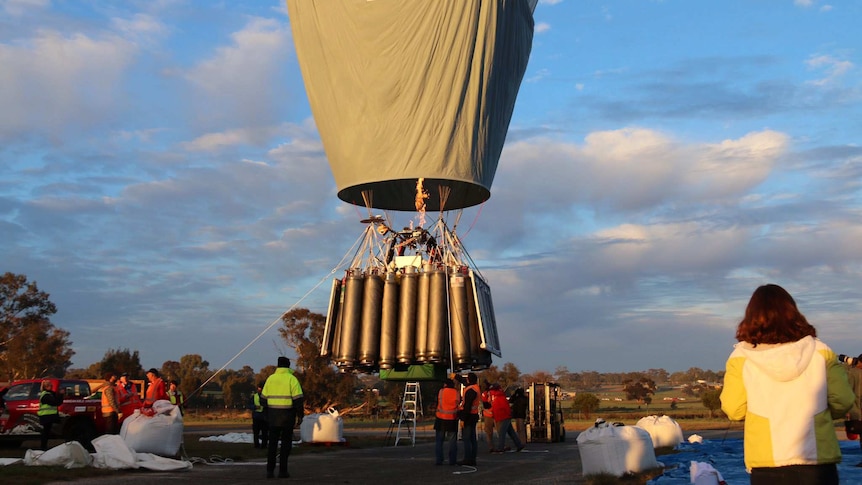A hot-air balloon inches away from the ground with oxygen bottles on the outside of the gondola as people watch on.