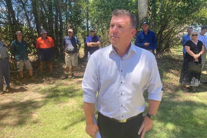 A man in a collared shirt stands in front of blue collar workers in a treed setting.