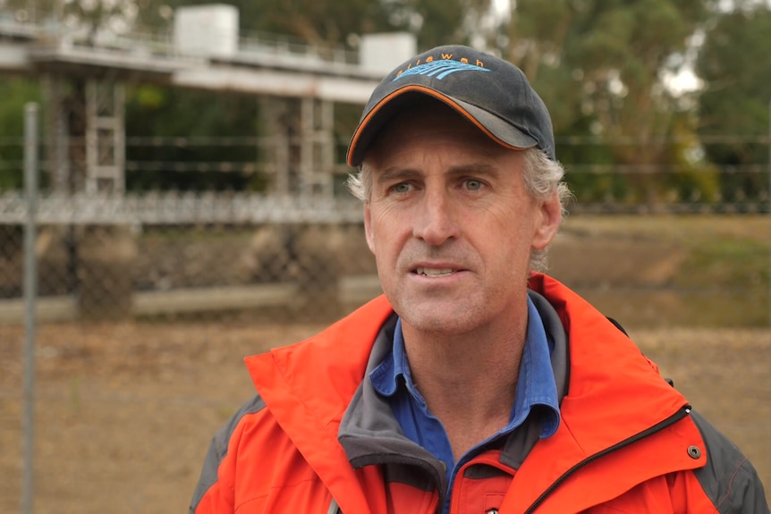 Man wearing cap and jacket with hi vis lining, standing in front of a fence along a river
