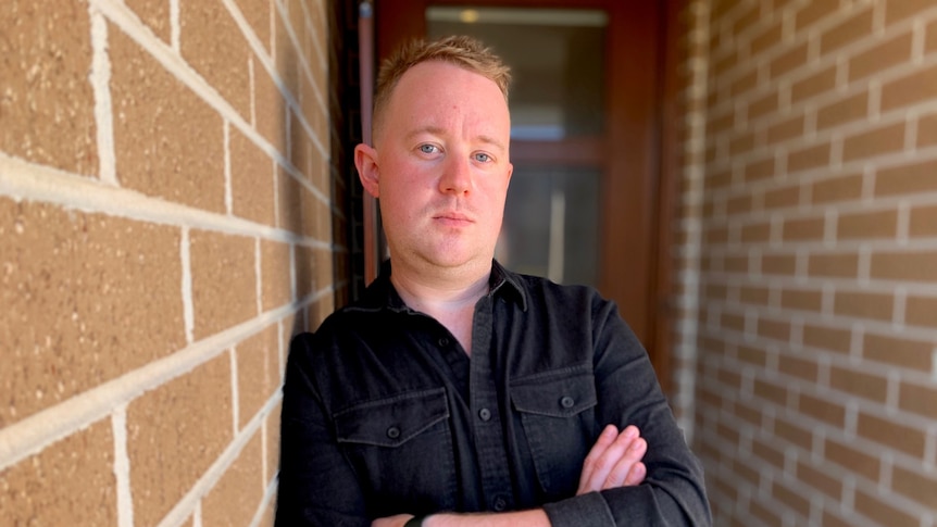 A man in a black shirt leans on a brick wall with his arms crossed.