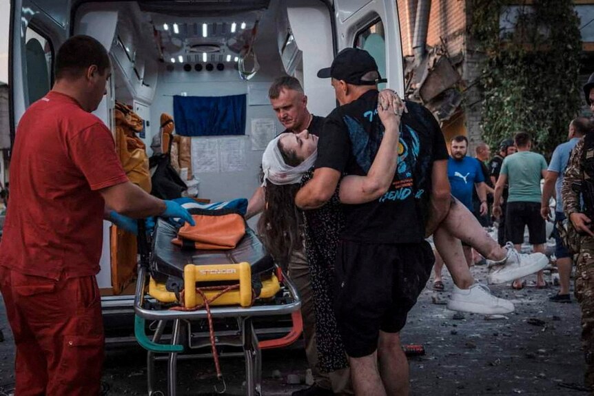 A woman with a bandage on her heard carried by rescuers to an ambulance stretcher at blast site.  