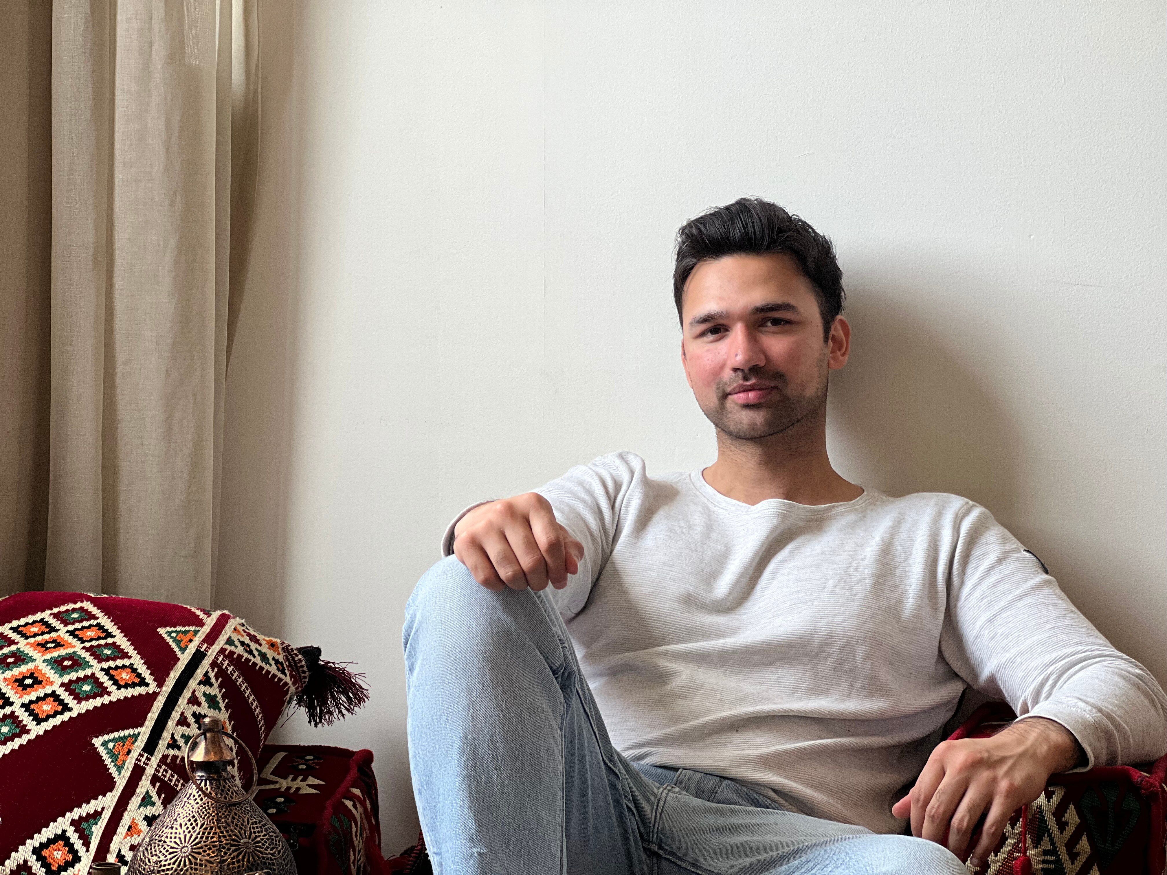 A young man sitting on the floor with one hand resting on his knee, the other on a red pillow. He's wearing a white top.