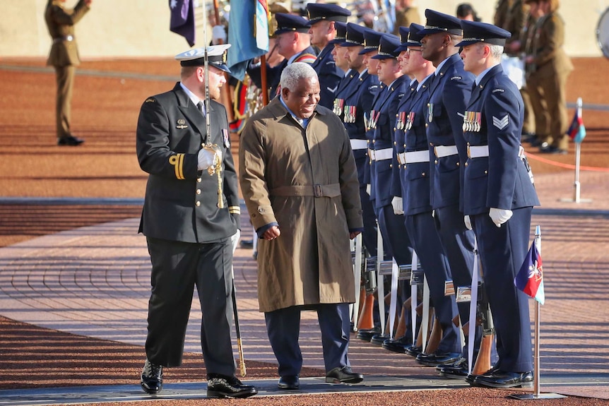 Rick Houenipwela, wearing a khaki coat, walks past a group of people in military dress uniforms.