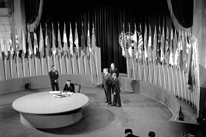 A black and white image of a man at a desk surrounded by national flags
