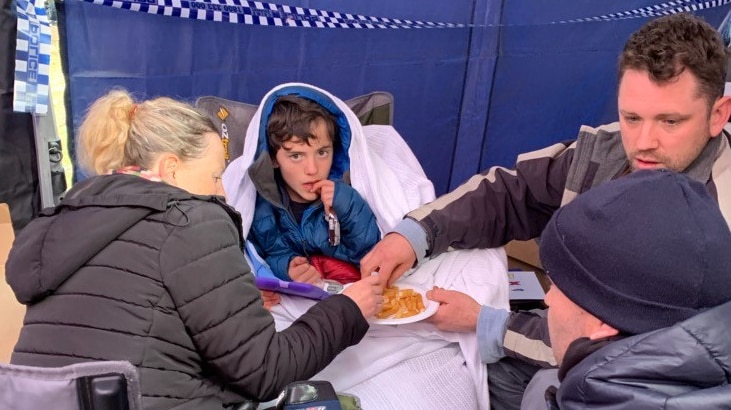 William Callaghan sits covered in a big jacket and blanket and eats hot chips with his family around him.