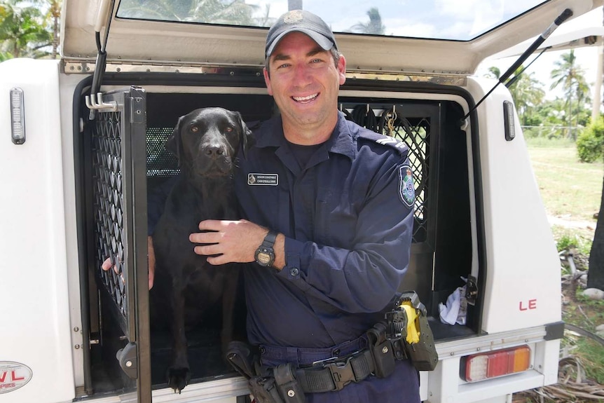 A black labrador is held by a police officer as they both sit at the back of a police vehicle
