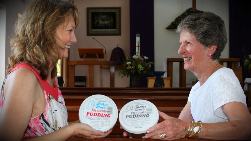 Two women sit at church pews inside a church holding packaged puddings labelled 'Father Mac's heavenly pudding'.