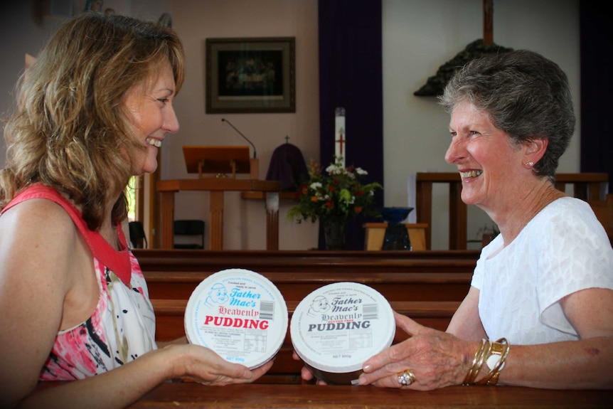 Two women sit at church pews inside a church holding packaged puddings labelled 'Father Mac's heavenly pudding'.