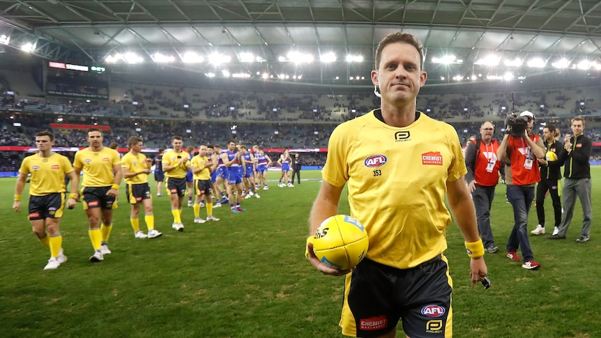 An AFL umpire faces the camera as he walks off the ground after a game, with other umpires and players following behind.