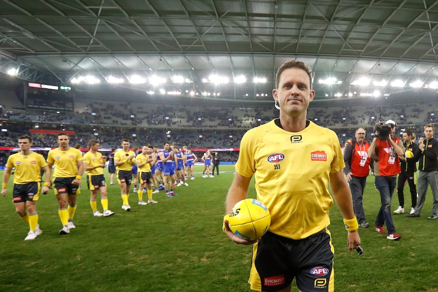 An AFL umpire faces the camera as he walks off the ground after a game, with other umpires and players following behind.