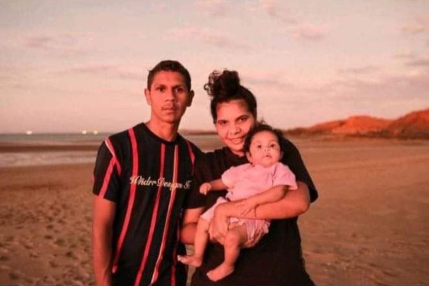 Man, woman holding little girl wearing pink standing on a beach.