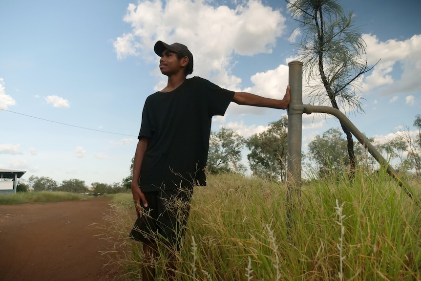 A boy with a brown cap and a black shirt stands on a dirt road, leaning on a fence post