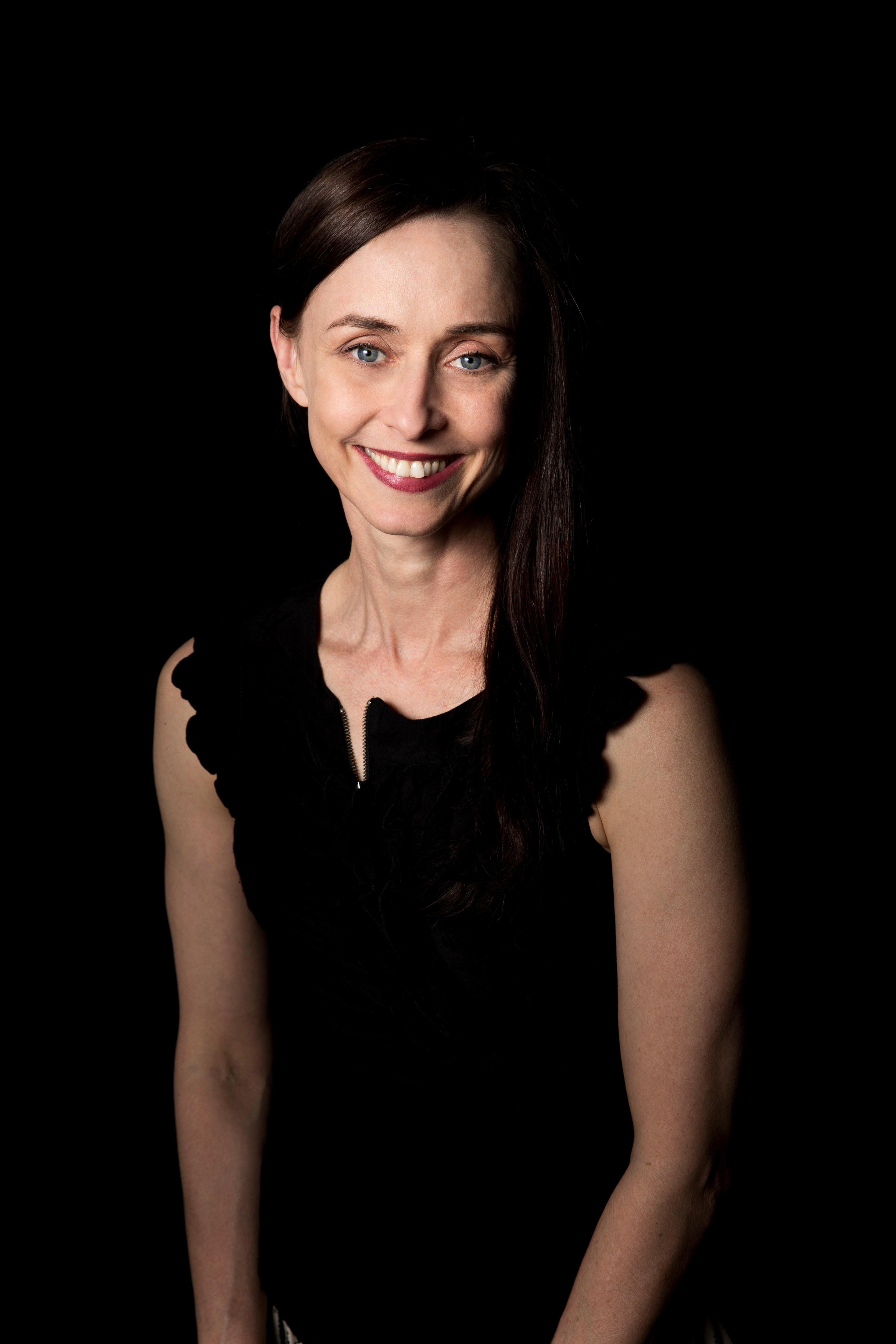 A headshot of a white woman in her 50s with mousey brown hair, wearing a black shirt and sitting against a black backdrop.