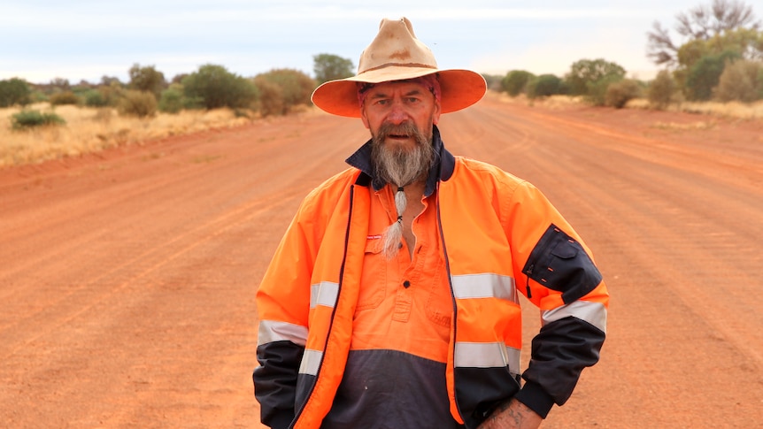 A man in fluro orange work gear and a wide brimmed hat, standing in the middle of a red dirt highway. 