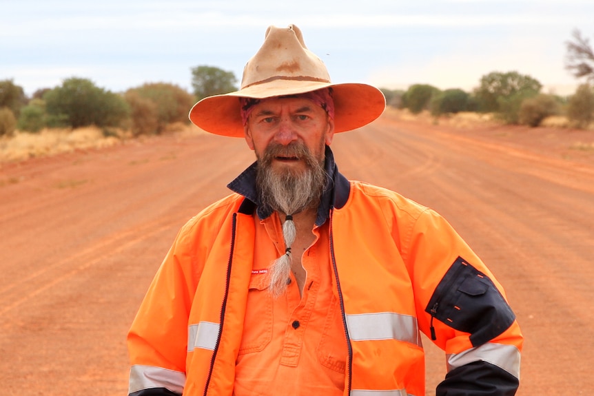 A man in fluro orange work gear and a wide brimmed hat, standing in the middle of a red dirt highway. 