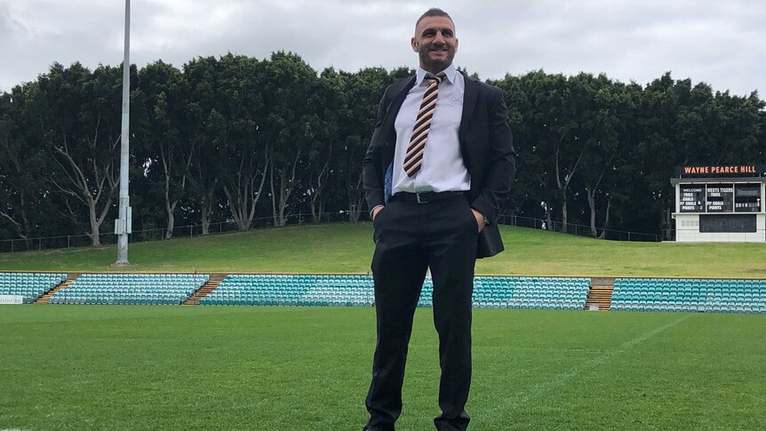 A man stands in a suit as he smiles while standing on a rugby league field.