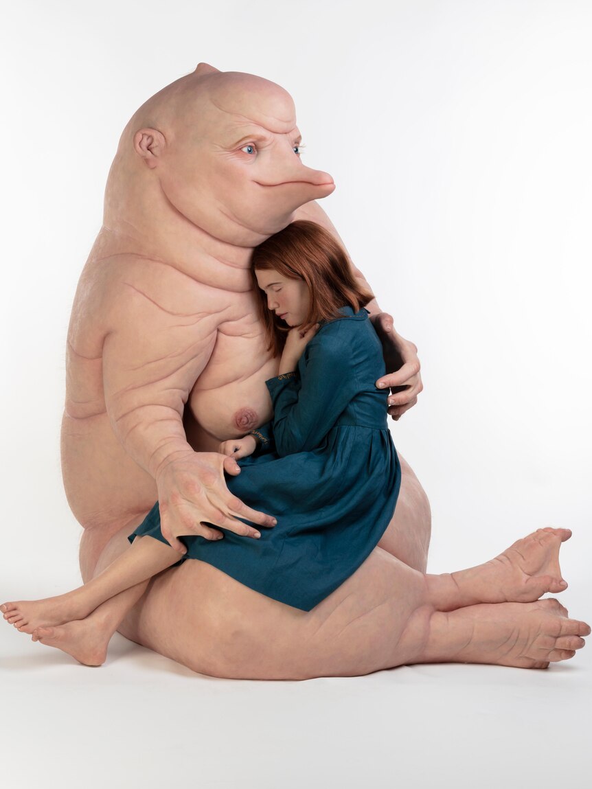 A large hyper-realistic sculpture of a humanlike creature cradling a woman in a blue dress.