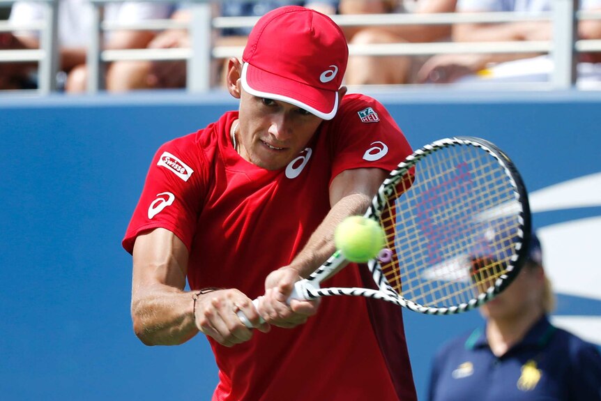 Alex de Minaur looks at the ball as he plays a double-handed shot wearing a red shirt and red cap