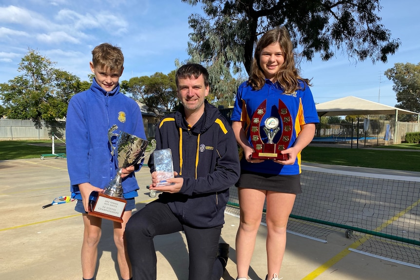 A boy, a man and girl pose with awards and trophies on a tennis court 