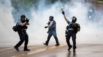 Police officers wearing gas masks walk in smoke as one throws a tear gas cannister.