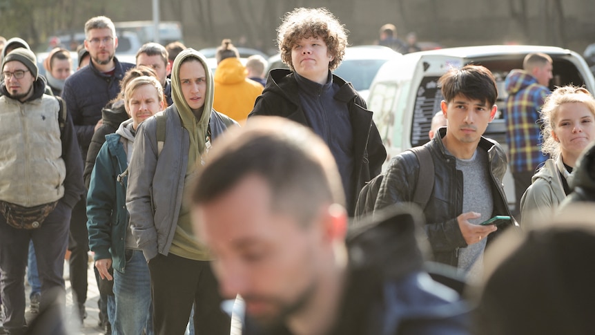 A group of people wearing jumpers and jackets line up on a sunny day.