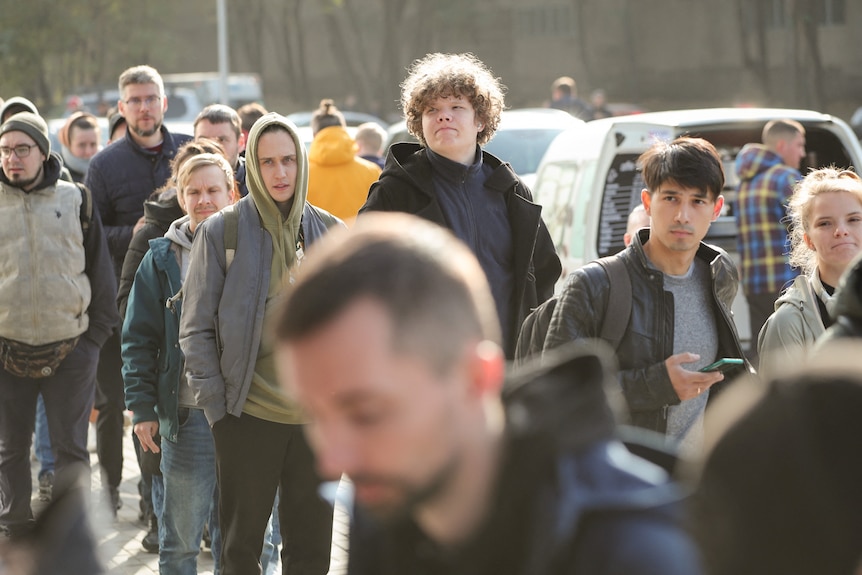 A group of people wearing jumpers and jackets line up on a sunny day.