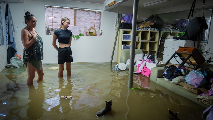 A woman and a girl wearing forlorn expressions stand in a flooded bedroom