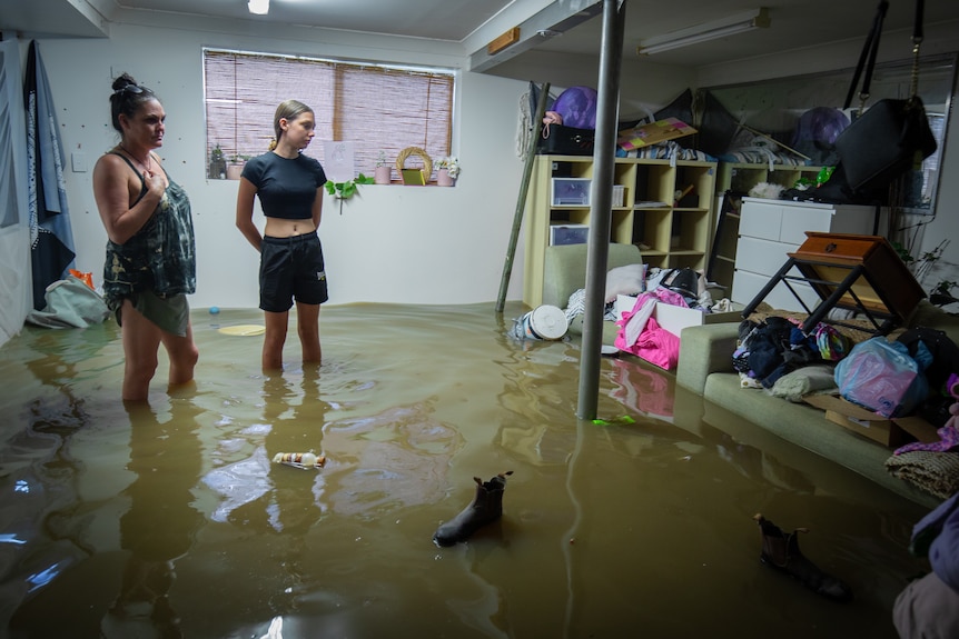 A woman and a girl wearing forlorn expressions stand in a flooded bedroom