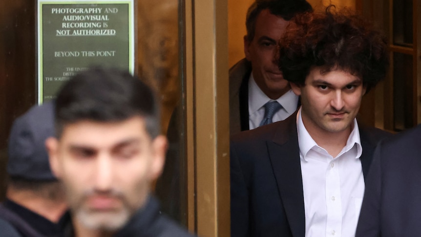 A young white man with frizzy dark hair looks at the camera as he exits a glass-doored building.