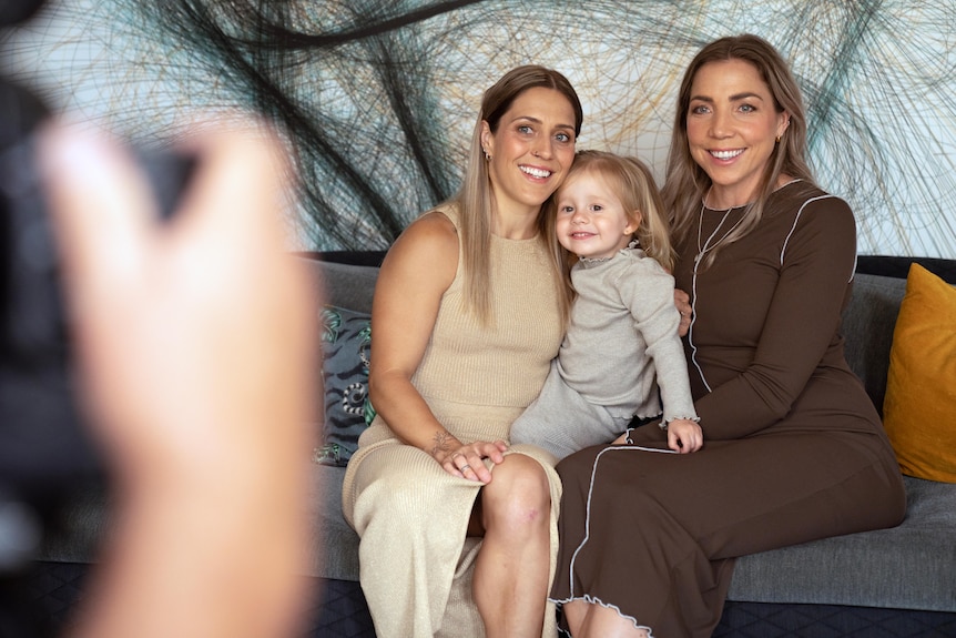 Two women seated on couch with toddler girl. The hands of a photographer are blurred in foreground