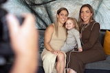 Two women seated on couch with toddler girl. The hands of a photographer are blurred in foreground