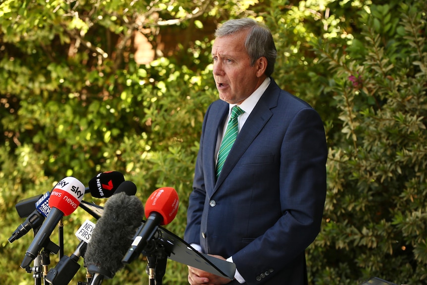 A wide shot of WA Environment Minister Reece Whitby speaking in a suit and tie outdoors, in front of microphones.