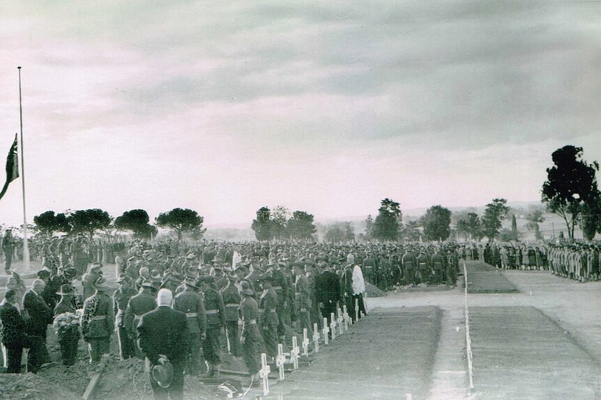 A black and white photograph of a military funeral service with many men in uniform below a flag pole at half mast.