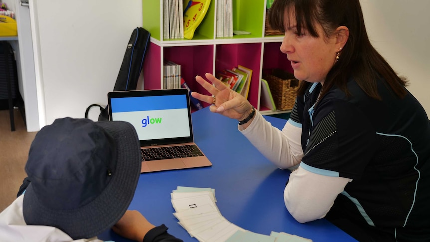 A female teacher sitting at a desk with a young boy, showing him a word.