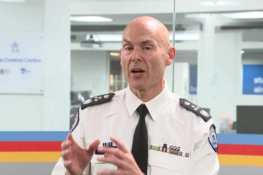 A bald man in a white uniform, black tie, talks to a crowd.