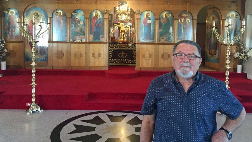 A man with white beard and wearing a blue shirt standing in front of a church alter