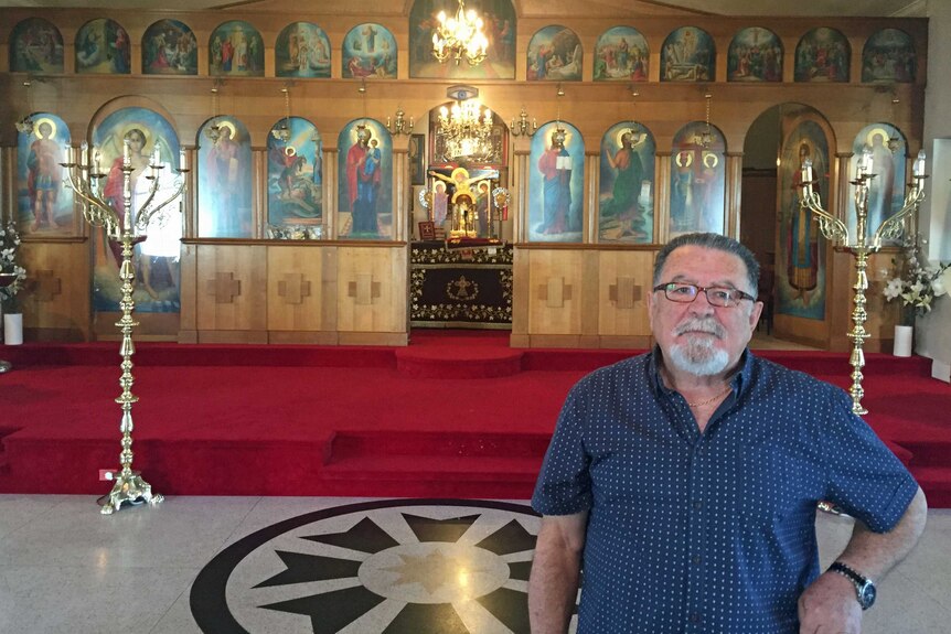 A man with white beard and wearing a blue shirt standing in front of a church alter