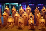 A group of monks in orange robes light candles in a darkened room.