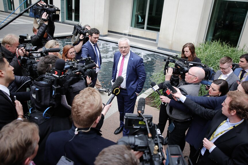 A media scrum surrounds a man with white hair wearing a navy suit in a courtyard next to a pond