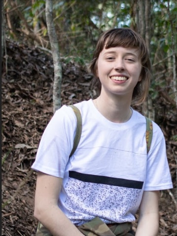 Emma sitting in a forest, she is smiling, surrounded by trees.