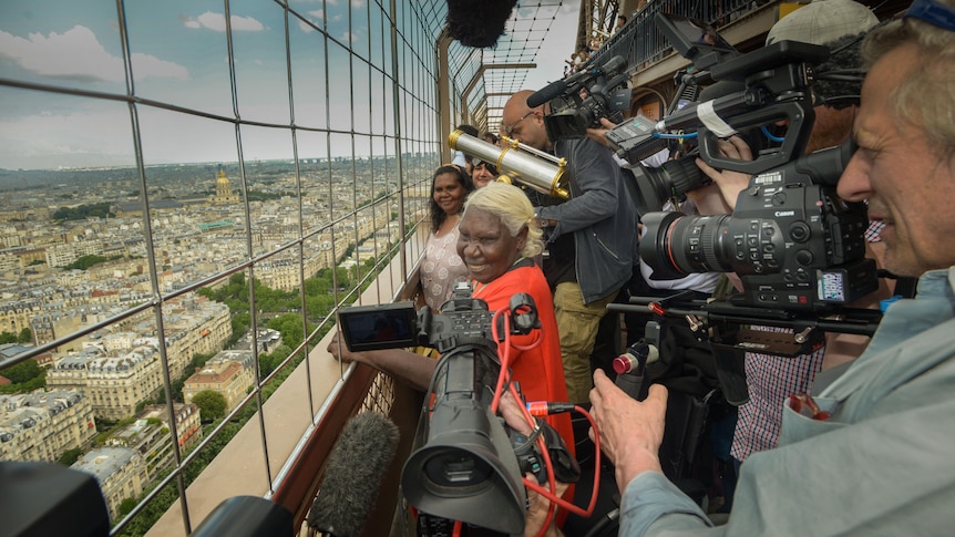 An Indigenous woman looks over the Paris skyline surrounded by tv cameras