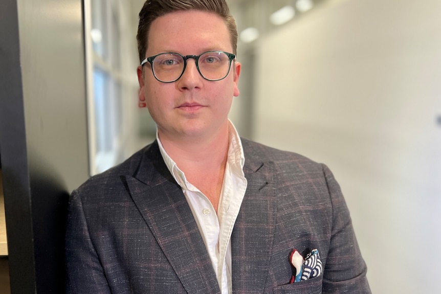 A man in a blazer and glasses looks at the camera.