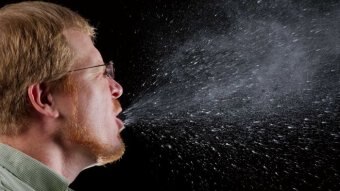 A man mid-sneeze against a black background, showing the salivary drops expelled from his mouth.