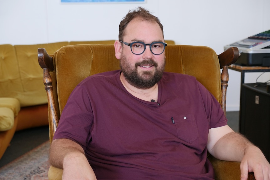 A man with brown hair and glasses, wearing a T-shirt, sits in a lounge chair. 