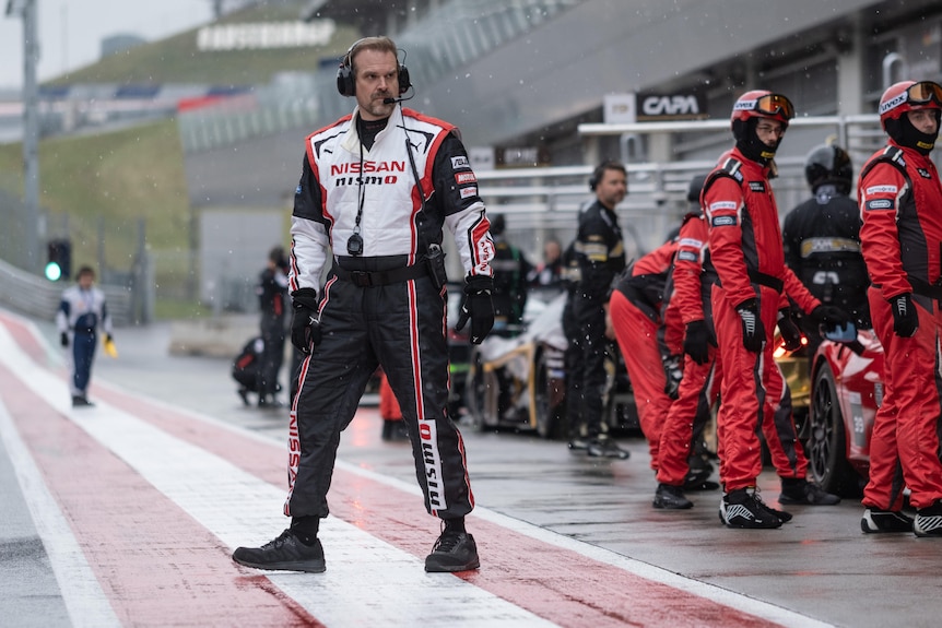 Actor David Harbour wearing a motor racing outfit with other race teams in the background.