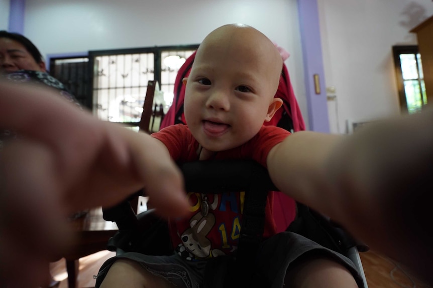 Baby Grammy reaches for the camera with both arms as he wears a red shirt.