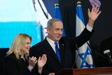 A suited man waves from a stage while standing next to a woman and Israeli flags.
